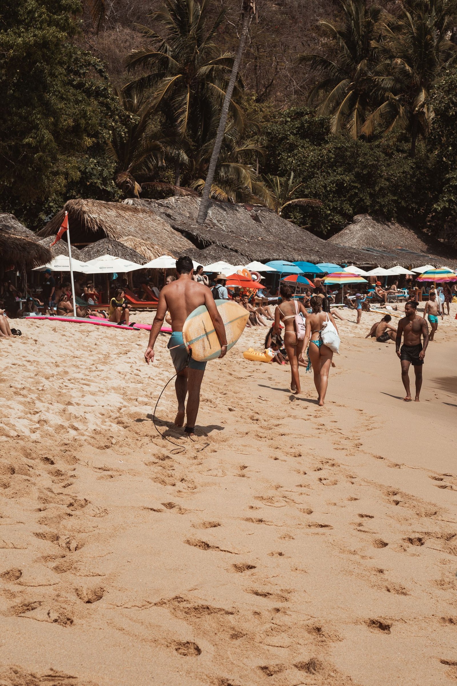 Crowded beach scene in Puerto Escondido with people and a surfer heading to the ocean.