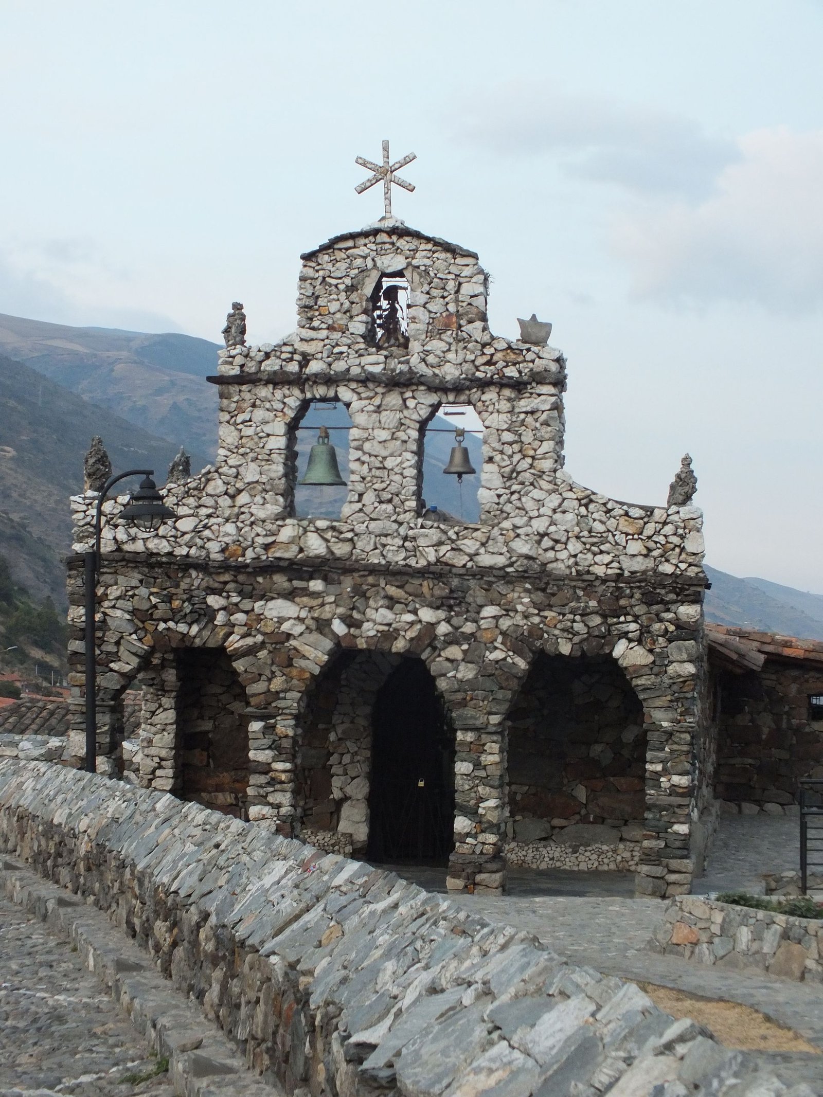 Rustic stone chapel with bells and cross at sunset in the mountains.
