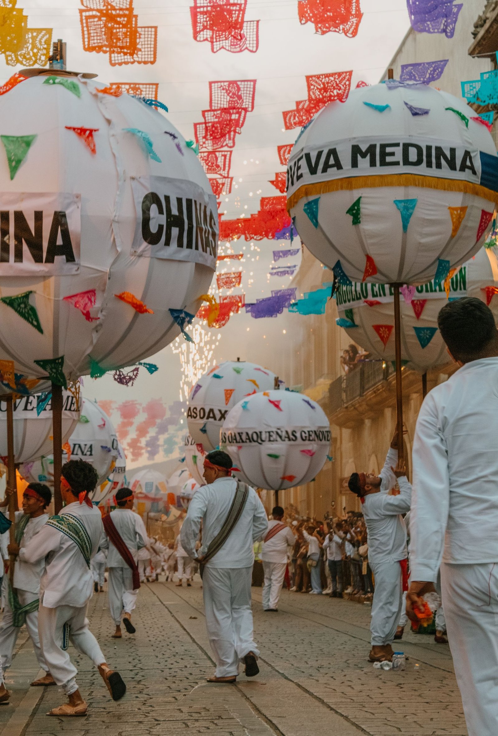 Vibrant street parade with people in traditional attire lifting decorated balloons during a festive celebration in Oaxaca.