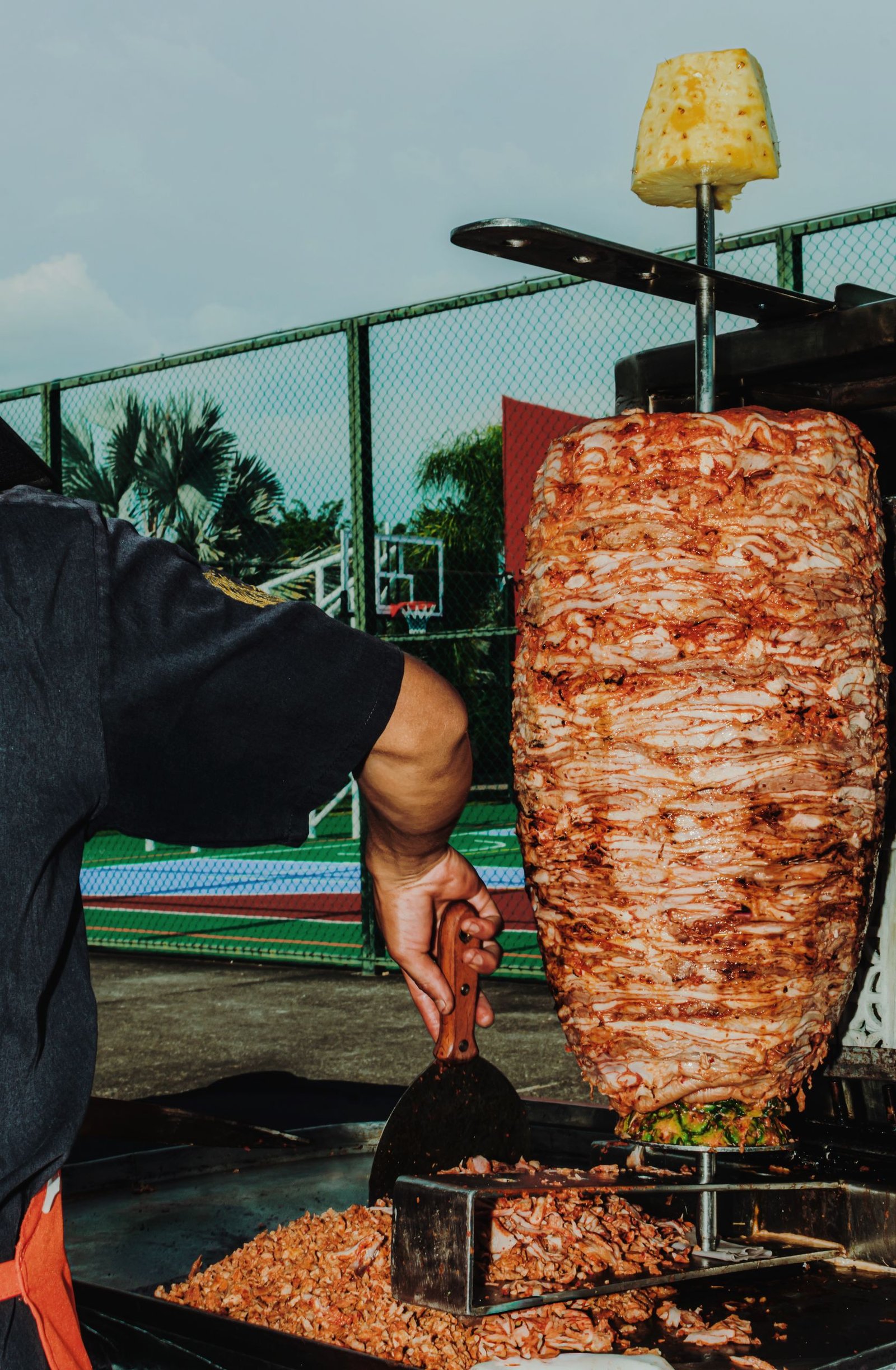 A vibrant scene of a street vendor slicing al pastor meat outdoors with a basketball court in the background.