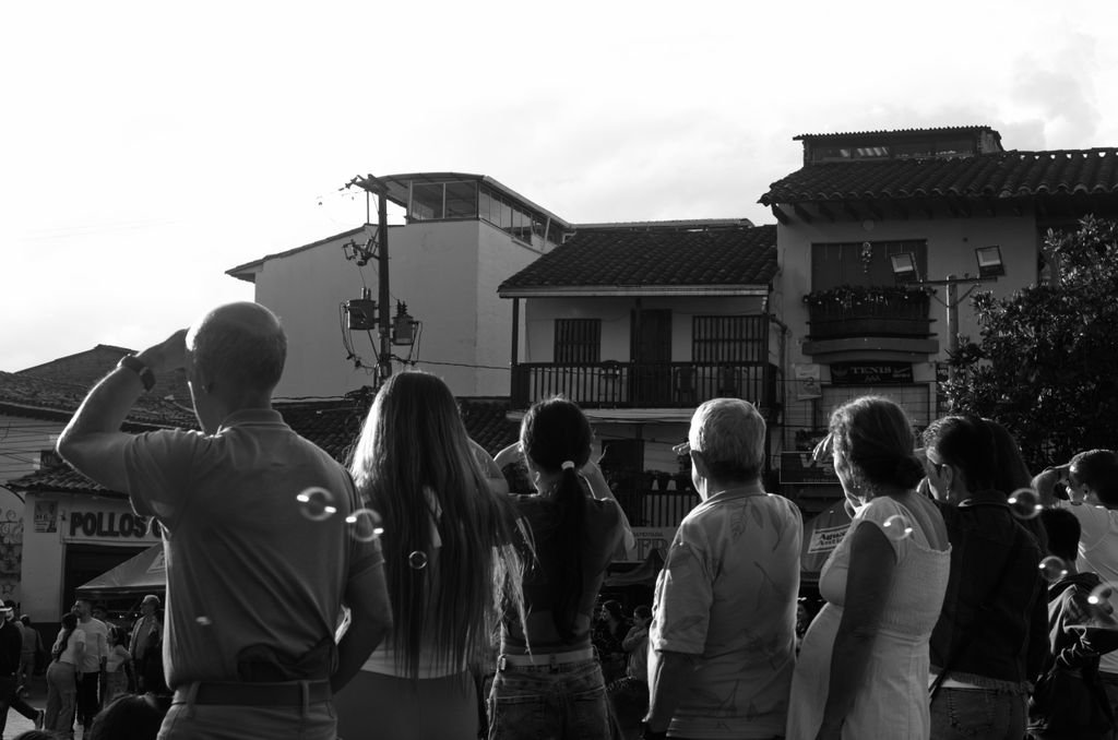 A diverse group of people watching a street event in a historic town captured in black and white.