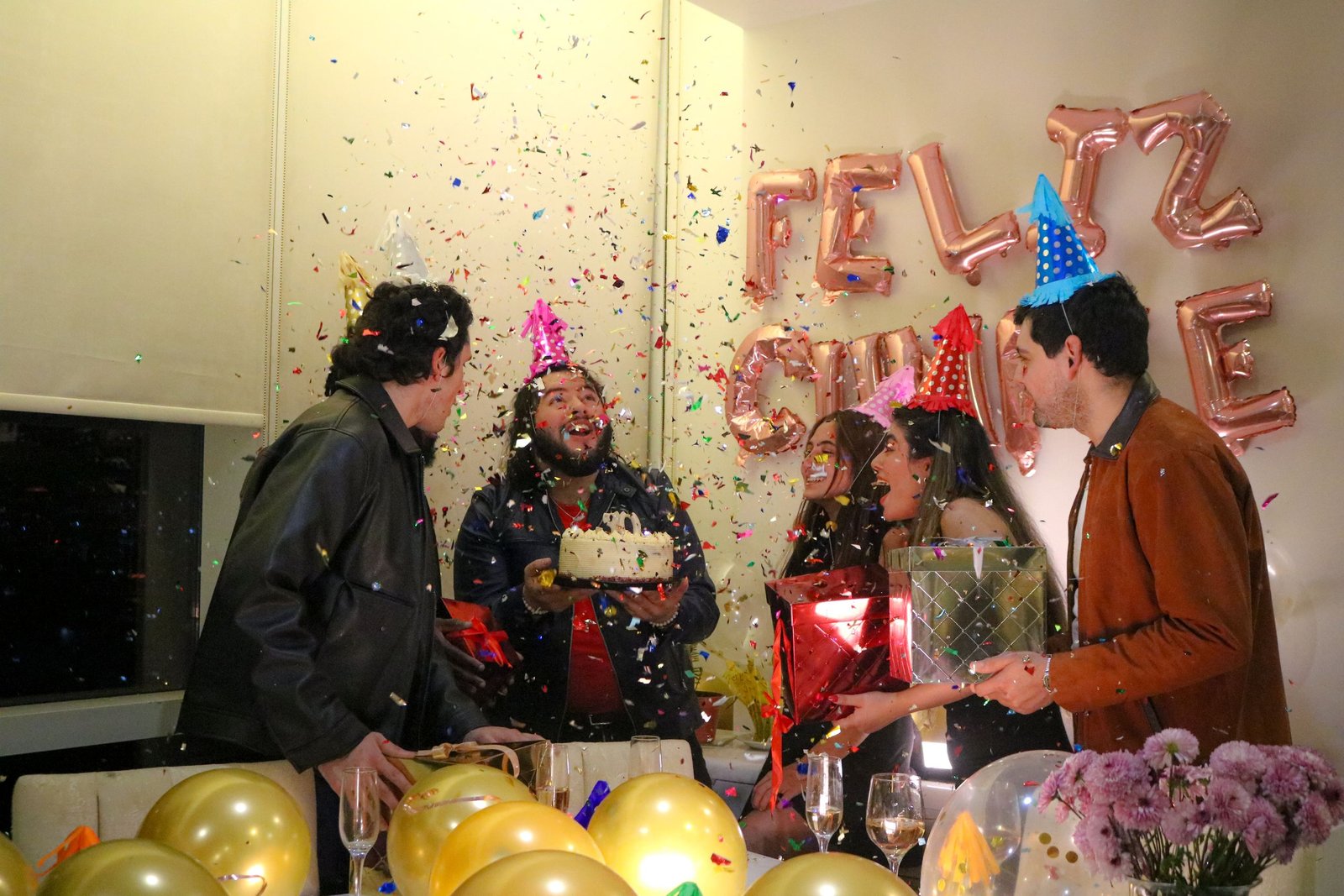 Group celebrating a birthday with cake, balloons, and confetti indoors.